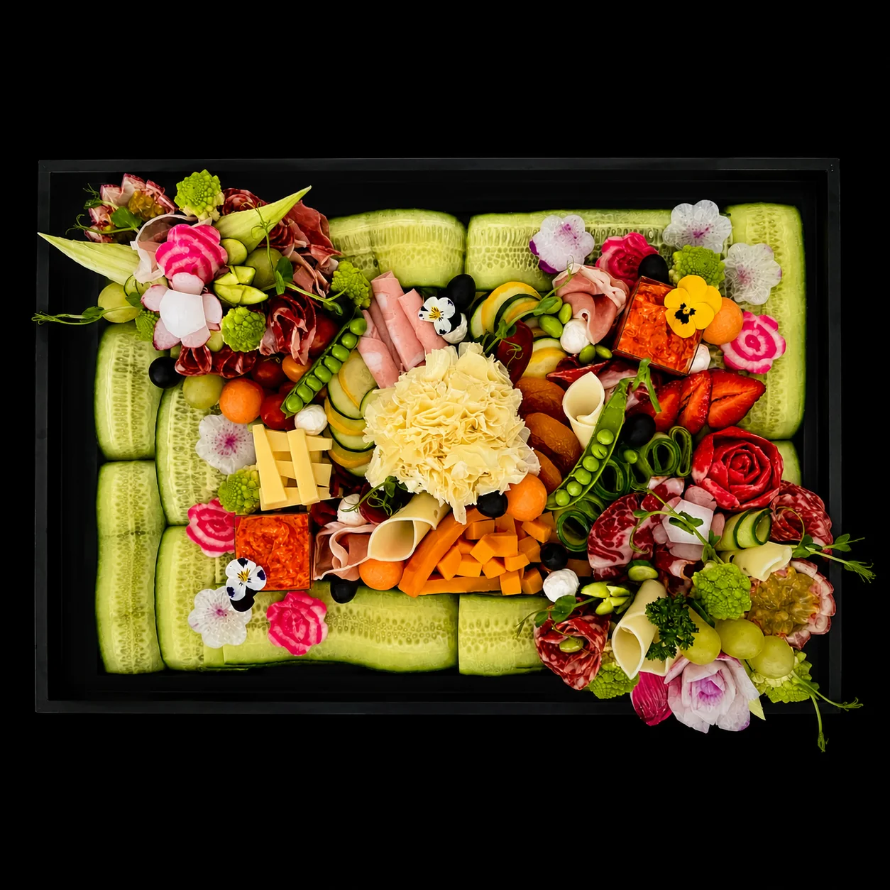 Plateau de charcuteries, fromages et fruits sculptés en roses, dressé sur un lit de concombre.