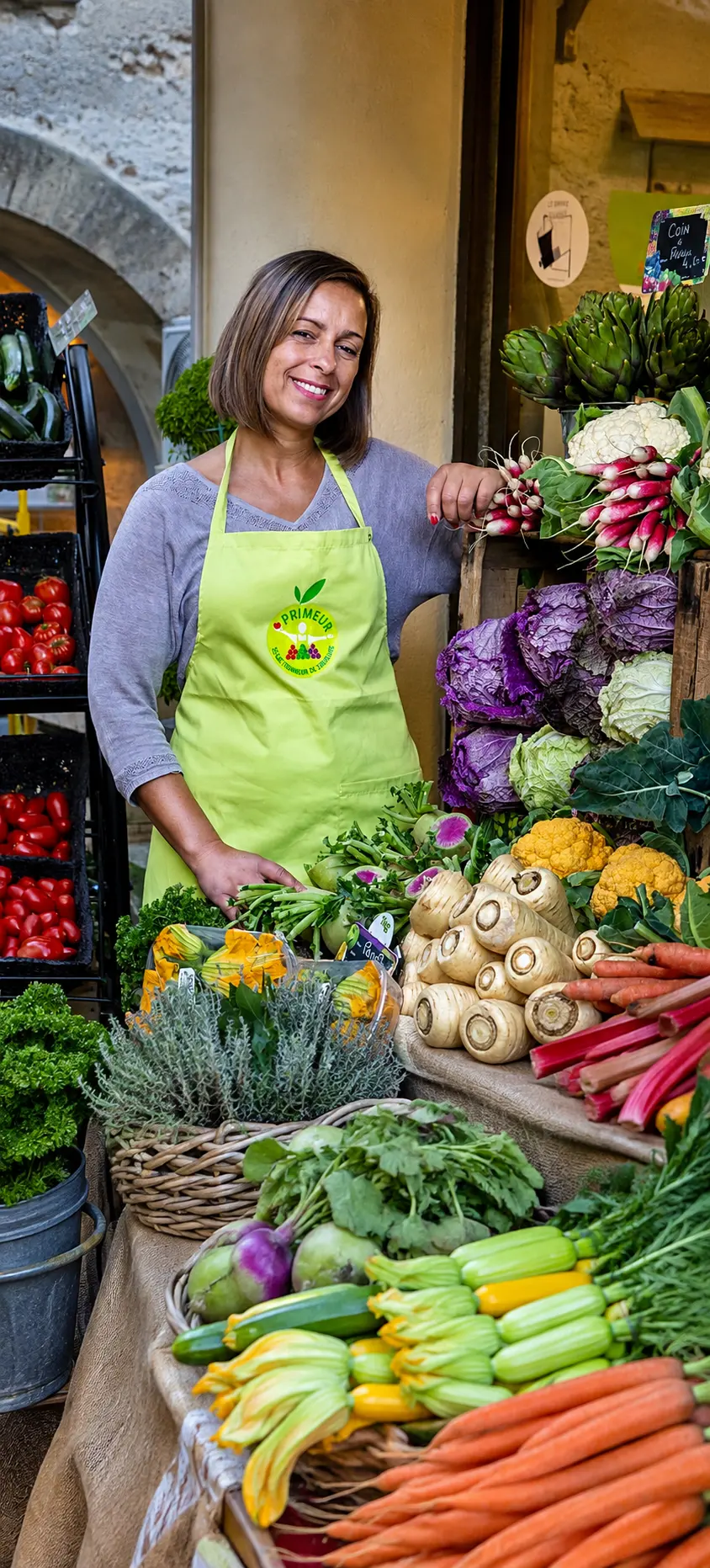 Samira Lemaire souriante derrière son étal, entourée de radis, choux et artichauts.