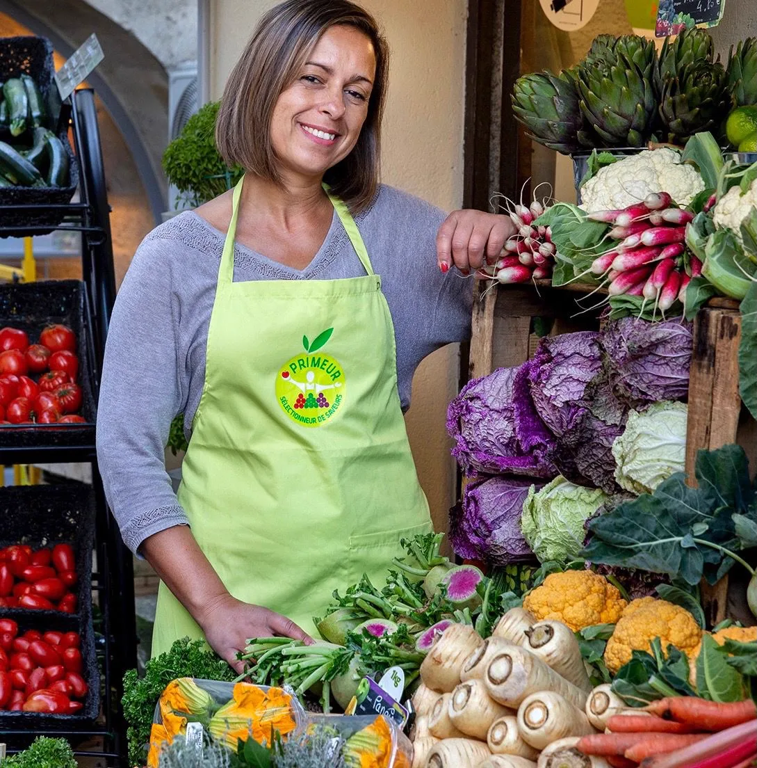 Samira Lemaire souriante derrière son étal, entourée de radis, choux et artichauts.
